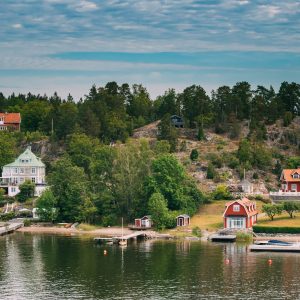Sweden. Many Beautiful Red Swedish Wooden Log Cabins Houses On Rocky Island Coast In Summer Sunny Evening. Lake Or River Landscape.